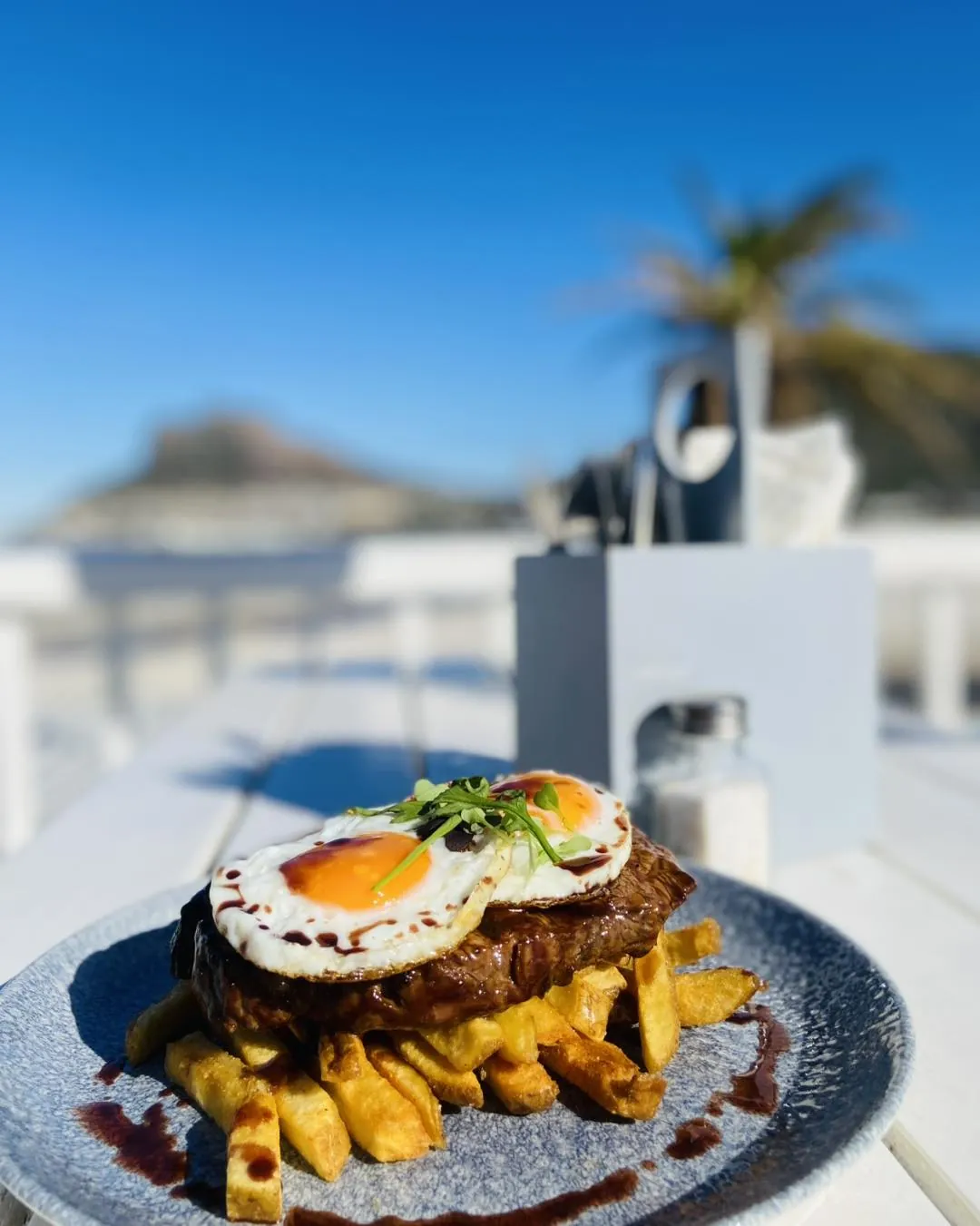 Steak, egg and chips on a table on the Dunes deck, with the bay and the Sentinel behind