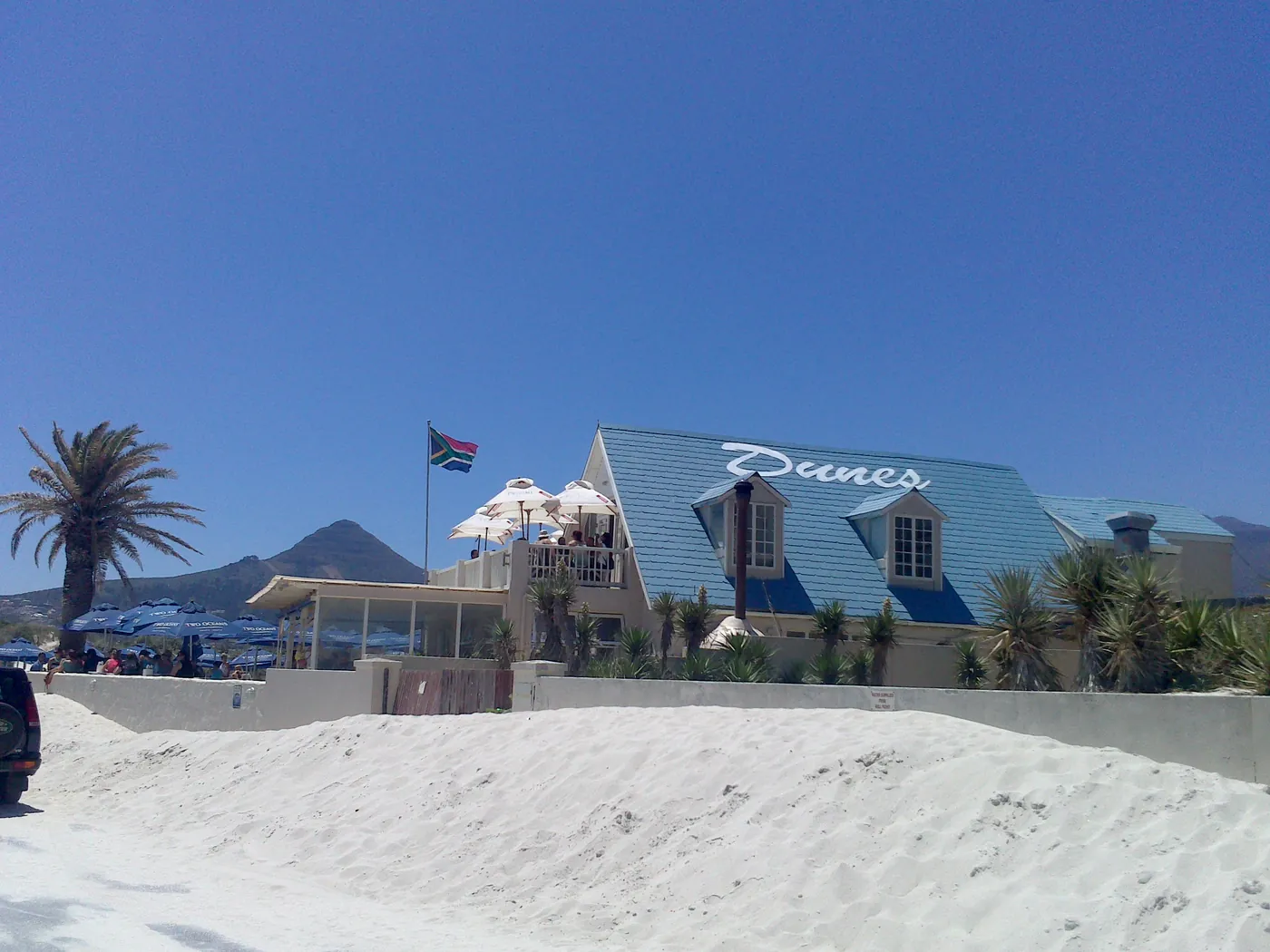 Dunes from the beach: the blue roof with the white Dunes script, palms, beach umbrellas and a sand dune in front